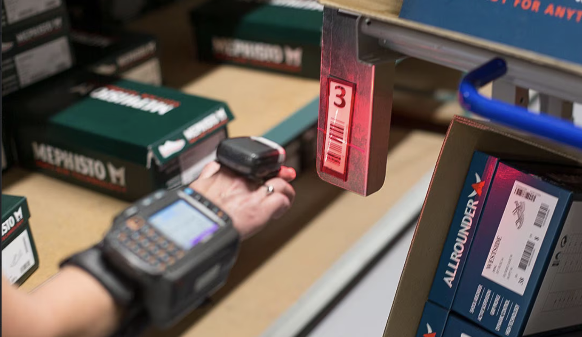 A worker wearing a ring scanner and wrist-mounted terminal, scanning a barcode on a shoe box on a warehouse shelf